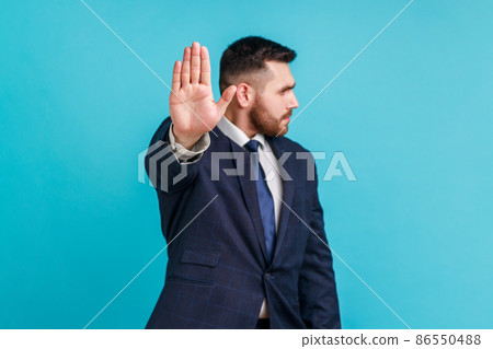Stop. keep your distance. Serious bearded man wearing official style suit standing with stop hand and looking away, showing ban, block sign with hand. Indoor studio shot isolated on blue background. 86550488