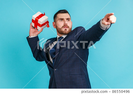 Catch your gift. Young serious businessman wearing dark official style suit, swung and wants to throw off your gift box, pointing finger in distance. Indoor studio shot isolated on blue background. Catch your gift. Young serious businessman wearing dark official style suit, swung and wants to throw off your gift box, pointing finger in distance. Indoor studio shot isolated on blue background. 86550496