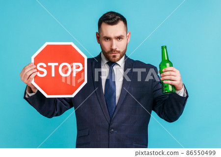 Stop drinking alcohol. Do not drive drunk. Anxious businessman showing alcoholic beverage beer bottle and stop sign, warning and worrying. Indoor studio shot isolated on blue background. 86550499