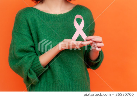 Female health check awareness. Woman hair in green casual style sweater holding pink ribbon, symbol of breast cancer, support of oncology patients. Indoor studio shot isolated on orange background. 86550700