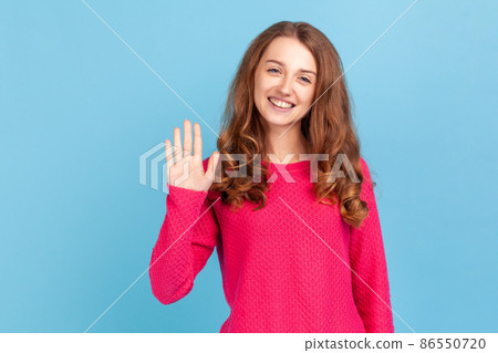 Attractive woman with wavy hair wearing pink pullover raised arm, saying hello, looking at camera with happy expression and toothy smile. Indoor studio shot isolated on blue background. 86550720