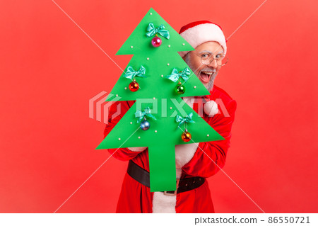 Amazed elderly man with gray beard wearing santa claus costume and sunglasses holding paper christmas tree looking at camera with excited expression. Indoor studio shot isolated on red background. 86550721