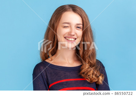 Positive woman with wavy hair wearing striped casual style sweater winking to camera, flirting expression, blinking eye as having cunning idea. Indoor studio shot isolated on blue background. 86550722