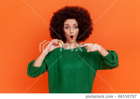 Portrait of surprised woman with curly hair wearing green casual style sweater holding and pointing at golden bitcoin, shocked with price. Indoor studio shot isolated on orange background. Portrait of surprised woman with curly hair wearing green casual style sweater holding and pointing at golden bitcoin, shocked with price. Indoor studio shot isolated on orange background. 86550787