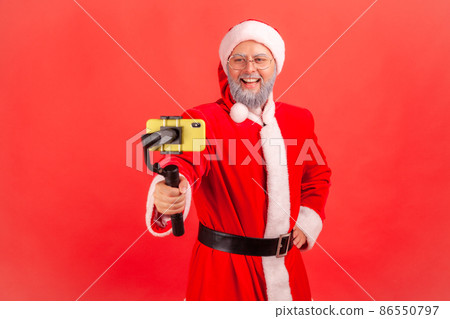 Attractive elderly man with gray beard wearing santa claus costume holding steadicam, broadcasting livestream, blogger talking with followers. Indoor studio shot isolated on red background. 86550797