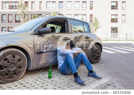 Man wearing jeans and blue shirt sitting near crashed car with bottle of alcohol beverage and sleeping, damaged automobile drunk driving, violation led to road accident. Outdoor shot. 86550806
