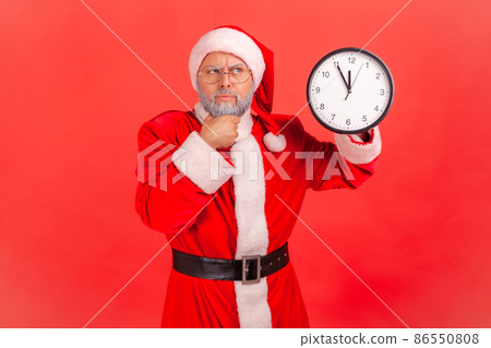 Pensive elderly man with gray beard wearing santa claus costume standing holding wall clock, holding chin, waiting Christmas party, looking away. Indoor studio shot isolated on red background. 86550808