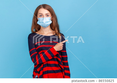 Portrait of serious woman wearing striped casual style sweater and surgical mask pointing finger side for empty wall for advertisement. Indoor studio shot isolated on blue background. 86550825