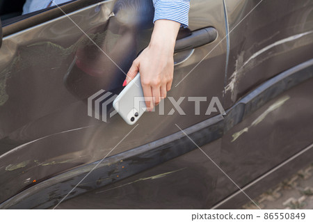 Closeup of woman hand with mobile phone hanging from automobile window, drunk driver in damaged car, injured female in automobile with dents and scratches, help for aggrieved. Outdoor shot. Closeup of woman hand with mobile phone hanging from automobile window, drunk driver in damaged car, injured female in automobile with dents and scratches, help for aggrieved. Outdoor shot. 86550849