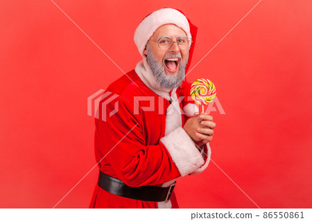 Portrait of happy amazed smiling elderly man with gray beard wearing santa claus costume standing with sugary lollypop in hands, looking at camera. Indoor studio shot isolated on red background. Portrait of happy amazed smiling elderly man with gray beard wearing santa claus costume standing with sugary lollypop in hands, looking at camera. Indoor studio shot isolated on red background. 86550861
