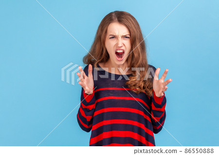 Angry woman with wavy hair wearing striped casual style sweater screaming frantically, proclaiming loud message, aggressive expression. Indoor studio shot isolated on blue background. 86550888