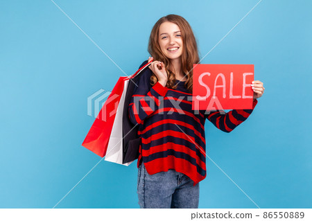 Cheerful woman wearing striped casual style sweater, boasting with purchases holding paper shopping bags and sale card, big holidays discounts. Indoor studio shot isolated on blue background. 86550889