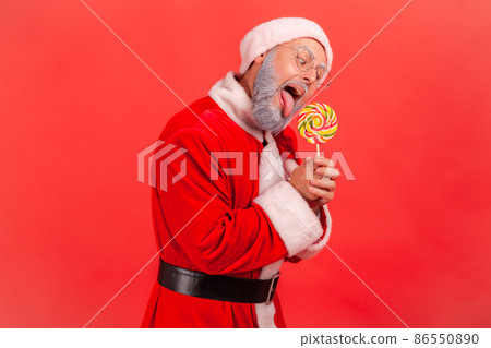 Side view of crazy hungry elderly man with gray beard wearing santa claus costume holding in hands sugary candy, biting multicolored lollipop. Indoor studio shot isolated on red background. Side view of crazy hungry elderly man with gray beard wearing santa claus costume holding in hands sugary candy, biting multicolored lollipop. Indoor studio shot isolated on red background. 86550890