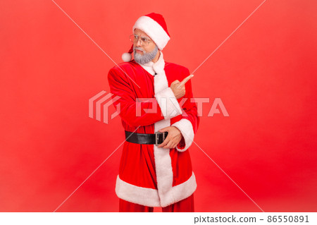 Annoyed elderly man with gray beard wearing santa claus costume showing way out with resentful irritated expression, ordering to leave, feeling betrayed. Indoor studio shot isolated on red background. 86550891