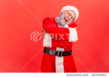 Elderly man with gray beard wearing santa claus costume hugging himself, feeling comfortable, narcissistic egoistic person, happy expression. Indoor studio shot isolated on red background. 86550892
