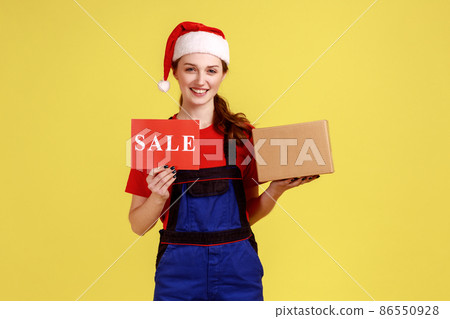Positive delivery woman with smile holding cardboard parcel and card with sale inscription, wearing blue overalls and santa claus hat. Indoor studio shot isolated on yellow background. Positive delivery woman with smile holding cardboard parcel and card with sale inscription, wearing blue overalls and santa claus hat. Indoor studio shot isolated on yellow background. 86550928