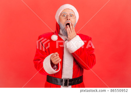 Astonished elderly man with gray beard wearing santa claus costume pointing to camera with finger, covering mouth with palm, hearing shocking news. Indoor studio shot isolated on red background. Astonished elderly man with gray beard wearing santa claus costume pointing to camera with finger, covering mouth with palm, hearing shocking news. Indoor studio shot isolated on red background. 86550952
