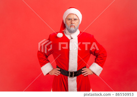 Confident elderly man with gray beard wearing santa claus costume standing with hands on hips and looking directly at camera with bossy look. Indoor studio shot isolated on red background. 86550953