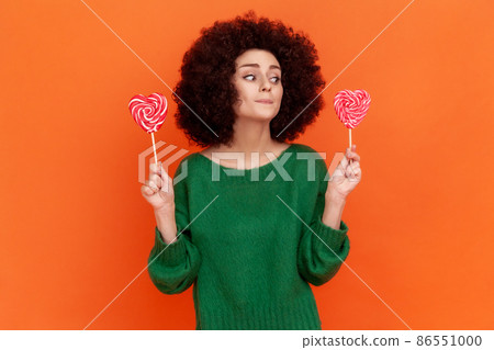 Attractive woman with Afro hairstyle wearing green casual style sweater keeping diet, decides eat candy or not, looking at sweets. Indoor studio shot isolated on orange background. Attractive woman with Afro hairstyle wearing green casual style sweater keeping diet, decides eat candy or not, looking at sweets. Indoor studio shot isolated on orange background. 86551000