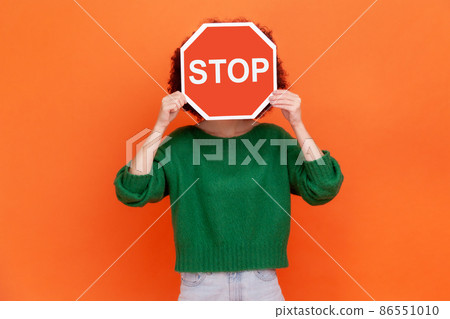 Unknown woman with wearing green casual style sweater hiding her face behind red stop traffic symbol, warning, avoids forbidden actions. Indoor studio shot isolated on orange background. Unknown woman with wearing green casual style sweater hiding her face behind red stop traffic symbol, warning, avoids forbidden actions. Indoor studio shot isolated on orange background. 86551010