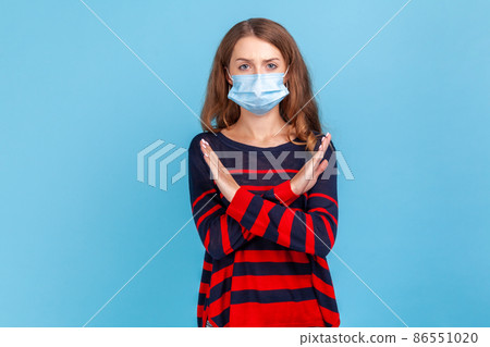 Portrait of worried woman wearing striped casual style sweater and surgical medical mask standing and showing closed or x sign at camera. Indoor studio shot isolated on blue background. 86551020