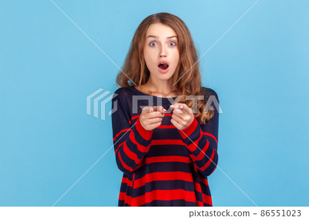 Astonished woman with wavy hair wearing striped casual style sweater pointing to camera with surprised shocked expression, making choice. Indoor studio shot isolated on blue background. Astonished woman with wavy hair wearing striped casual style sweater pointing to camera with surprised shocked expression, making choice. Indoor studio shot isolated on blue background. 86551023