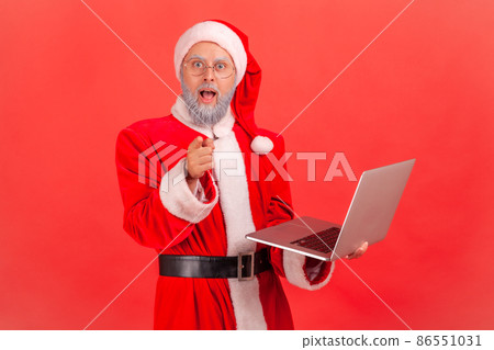 Shocked elderly man with gray beard wearing santa claus costume holding laptop in hands and pointing to camera with big eyes and astonishment. Indoor studio shot isolated on red background. 86551031