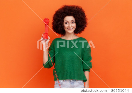 Portrait of smiling woman with curly hair wearing green casual style sweater standing holding red retro land line phone, waiting conversation. Indoor studio shot isolated on orange background. 86551044