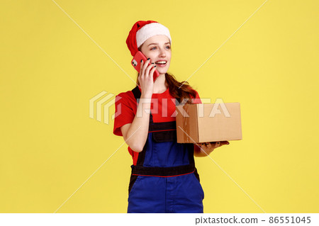 Friendly delivery woman talking on cellphone with client and accepting order, holding cardboard box, wearing blue overalls and santa claus hat. Indoor studio shot isolated on yellow background. Friendly delivery woman talking on cellphone with client and accepting order, holding cardboard box, wearing blue overalls and santa claus hat. Indoor studio shot isolated on yellow background. 86551045