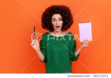 Surprised woman with Afro hairstyle wearing green casual style sweater standing with open mouth, holding paper notebook and pencil. Indoor studio shot isolated on orange background. 86551052