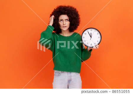 Portrait of pensive woman with curly hair wearing green casual style sweater holding wall clock and scratching her head, deadline, punctuality. Indoor studio shot isolated on orange background. Portrait of pensive woman with curly hair wearing green casual style sweater holding wall clock and scratching her head, deadline, punctuality. Indoor studio shot isolated on orange background. 86551055