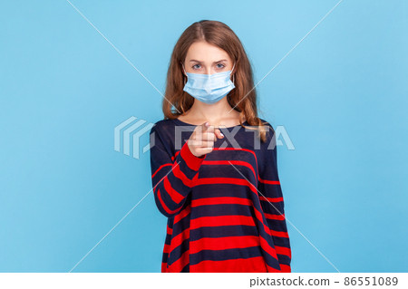 Hey you. take care of you health. Portrait of serious woman wearing sweater and surgical medical mask standing, pointing and looking at camera and blame. Indoor studio shot isolated on blue background Hey you. take care of you health. Portrait of serious woman wearing sweater and surgical medical mask standing, pointing and looking at camera and blame. Indoor studio shot isolated on blue background 86551089