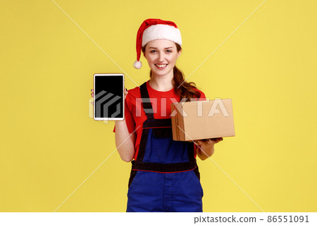 Positive courier female holding cardboard parcel and tablet with empty display for advertisement, wearing blue overalls and santa claus hat. Indoor studio shot isolated on yellow background. Positive courier female holding cardboard parcel and tablet with empty display for advertisement, wearing blue overalls and santa claus hat. Indoor studio shot isolated on yellow background. 86551091