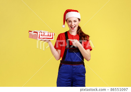Excited delivery woman holding wrapped present box on palm and pointing at gift with finger, wearing blue overalls and santa claus hat. Indoor studio shot isolated on yellow background. Excited delivery woman holding wrapped present box on palm and pointing at gift with finger, wearing blue overalls and santa claus hat. Indoor studio shot isolated on yellow background. 86551097