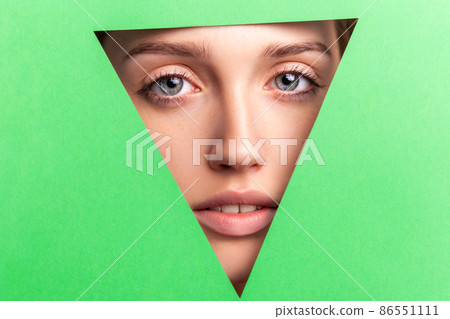 Closeup portrait of well-groomed woman with natural makeup, looking at camera through triangular hole of green paper, beauty and health and skin care concept. Indoor studio shot. 86551111
