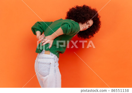 Young adult woman with Afro hairstyle wearing green casual style sweater suffering from lower back pain, pinched nerve or kidney inflammation. Indoor studio shot isolated on orange background. Young adult woman with Afro hairstyle wearing green casual style sweater suffering from lower back pain, pinched nerve or kidney inflammation. Indoor studio shot isolated on orange background. 86551138