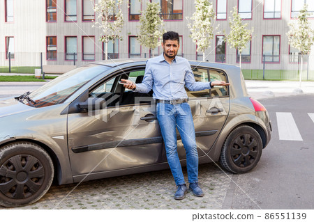 Young adult bearded man with sad expression standing with raised arms and looking at camera, being confused after car accident, broken auto with dents and scratches on side part. Outdoor shot. Young adult bearded man with sad expression standing with raised arms and looking at camera, being confused after car accident, broken auto with dents and scratches on side part. Outdoor shot. 86551139