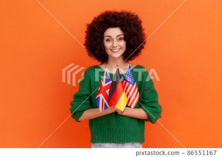 Portrait of beautiful woman with Afro hairstyle wearing green casual style sweater holding in hands British, German and American flags. Indoor studio shot isolated on orange background. 86551167