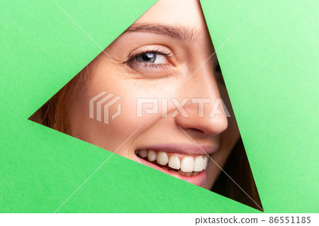 Young attractive woman in triangle with beautiful eyes and charming smile looking into the hole of green paper. Beauty, makeup health and skin care concept. Indoor studio shot. Young attractive woman in triangle with beautiful eyes and charming smile looking into the hole of green paper. Beauty, makeup health and skin care concept. Indoor studio shot. 86551185