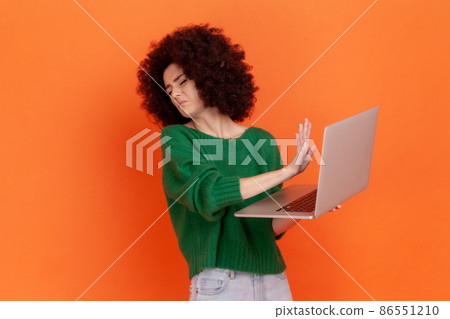 Portrait of woman with Afro hairstyle wearing green casual style sweater turning face from laptop display and showing stop gesture, forbidden content. Indoor studio shot isolated on orange background. Portrait of woman with Afro hairstyle wearing green casual style sweater turning face from laptop display and showing stop gesture, forbidden content. Indoor studio shot isolated on orange background. 86551210