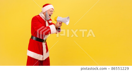 Side view of angry screaming elderly man with gray beard wearing santa claus costume holding megaphone and yelling with aggressive expression. Indoor studio shot isolated on yellow background. 86551229