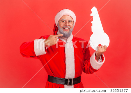 Portrait of smiling elderly man with gray beard wearing santa claus costume standing pointing to paper key, recommend agency for buying apartment. Indoor studio shot isolated on red background. Portrait of smiling elderly man with gray beard wearing santa claus costume standing pointing to paper key, recommend agency for buying apartment. Indoor studio shot isolated on red background. 86551277
