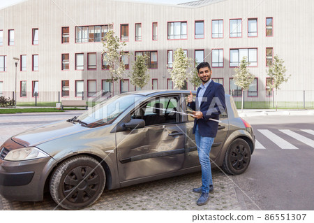 Portrait of young adult bearded insurance agent standing with positive facial expression near wrecked car and showing thumb up, wearing jeans and jacket, examining auto after accident. Outdoor shot. Portrait of young adult bearded insurance agent standing with positive facial expression near wrecked car and showing thumb up, wearing jeans and jacket, examining auto after accident. Outdoor shot. 86551307