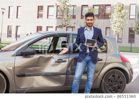 Young adult bearded man working in insurance service posing near dented automobile, holding tablet with empty display for advertisement and showing dents and scratches on car door. Outdoor shot. 86551360