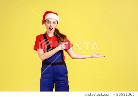 Female worker presenting copy space on palm, points finger aside, advertisement of service industry, wearing blue overalls and santa claus hat. Indoor studio shot isolated on yellow background. Female worker presenting copy space on palm, points finger aside, advertisement of service industry, wearing blue overalls and santa claus hat. Indoor studio shot isolated on yellow background. 86551365