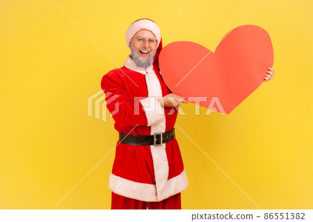 Satisfied elderly man with gray beard wearing santa claus costume holding red heart in hands, expressing sensitive feelings, looking at camera. Indoor studio shot isolated on yellow background. Satisfied elderly man with gray beard wearing santa claus costume holding red heart in hands, expressing sensitive feelings, looking at camera. Indoor studio shot isolated on yellow background. 86551382