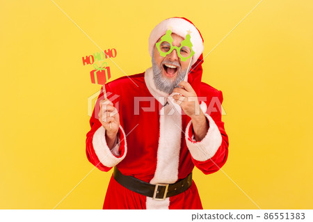 Excited elderly man with gray beard wearing santa claus costume holding congratulations cards on sticks, new year and christmas celebration. Indoor studio shot isolated on yellow background. 86551383