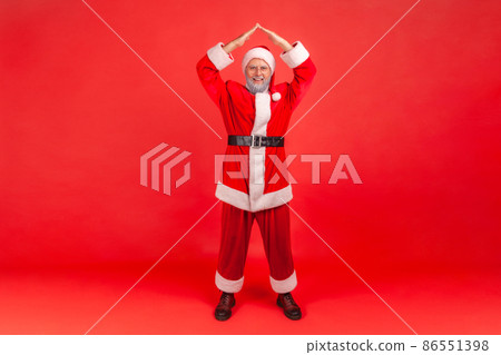Full length of elderly man with gray beard wearing santa claus costume making roof of hands above head looking at camera, feeling himself in safety. Indoor studio shot isolated on red background. Full length of elderly man with gray beard wearing santa claus costume making roof of hands above head looking at camera, feeling himself in safety. Indoor studio shot isolated on red background. 86551398