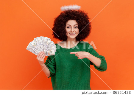 Young adult angelic woman with curly hair wearing green casual style sweater with nimb over head holding and pointing at fan of dollars banknotes. Indoor studio shot isolated on orange background. 86551428