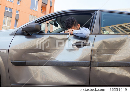Despaired man sitting behind the wheel of crashed car and puts his head down, crying, broke his car due to traffic violence, scratches and dents on auto door. Outdoor shot. Despaired man sitting behind the wheel of crashed car and puts his head down, crying, broke his car due to traffic violence, scratches and dents on auto door. Outdoor shot. 86551449